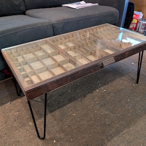 May include: A rectangular coffee table with a glass top, showcasing a vintage printer's tray interior. The table has a dark brown wooden frame and is supported by black hairpin legs. The interior is divided into numerous small compartments.