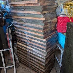 May include: A tall stack of vintage wooden drawers, likely from a printing press or similar industrial equipment. The drawers are a mix of brown and olive green, with metal handles. The stack is on wheels.