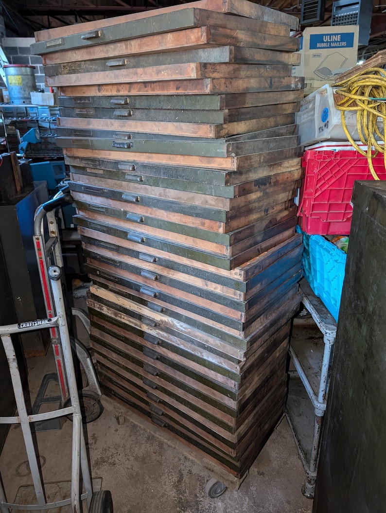 May include: A tall stack of vintage wooden drawers, likely from a printing press or similar industrial equipment. The drawers are a mix of brown and olive green, with metal handles. The stack is on wheels.