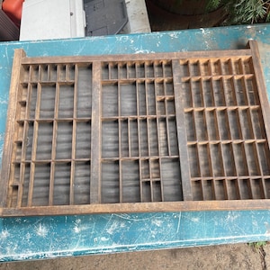 May include: A rectangular wooden printer's type case featuring a grid of numerous small, open compartments of varying sizes. The wood has a weathered, dark brown finish, and the interior base of each section is a dark, textured material.