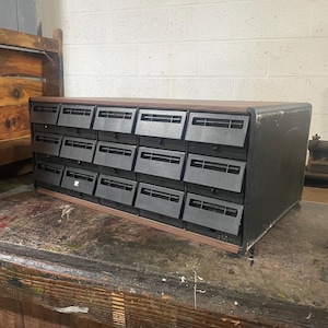 May include: A rectangular storage cabinet featuring fifteen black plastic drawers arranged in three rows of five. The unit has a dark brown top surface and black side panels, with each drawer front including a horizontal slot for a label.