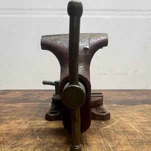 May include: A red metal vise with a long, dark metal handle. The vise is sitting on a wooden surface.