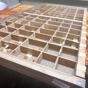 Puede incluir: Una bandeja de tipógrafo de madera vintage con numerosos compartimentos pequeños, probablemente utilizada para organizar tipos u objetos pequeños. La bandeja está hecha de madera de color claro y sujeta con cinta adhesiva. Una etiqueta con el número 18910 es visible.
