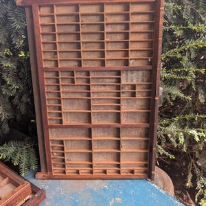 May include: A well-used wooden printer's tray, featuring a grid of small compartments. The tray is constructed from dark brown wood and has a rectangular form. The compartments are of various sizes, suggesting it was used for storing small items or type.