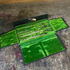May include: A vintage drafting instrument set housed in a black folding case with a vibrant green velvet interior. The set includes several metal compasses and dividers secured in molded slots, with a worn label on the inner lid.