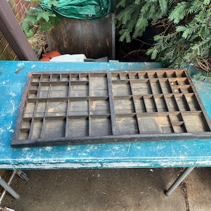 May include: A rectangular wooden printer's type drawer featuring a dark brown finish and numerous small, varied compartments arranged in a grid pattern. The wooden tray has visible signs of wear and age, with open slots for organizing small items.