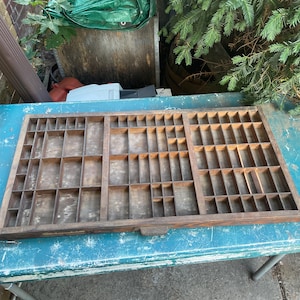May include: A rectangular wooden printer's drawer featuring a grid of numerous small, open compartments of varying sizes. The wood has a weathered, dark brown finish with visible grain and signs of age throughout the interior and exterior frame.