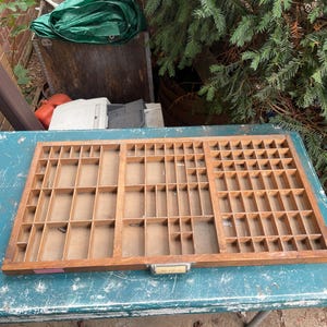 May include: A rectangular wooden printer's type drawer featuring numerous small, varied compartments for organising items. The light brown wood frame includes a metal label holder at the bottom centre with a small, faded paper insert inside.