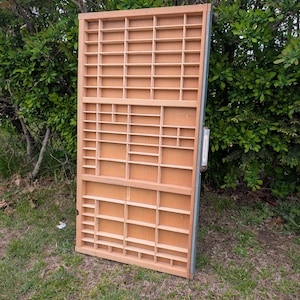 May include: A large, rectangular wooden display case with numerous small compartments. The case is made of light-colored wood and has a grid-like structure. The case is leaning against a backdrop of green foliage and grass.