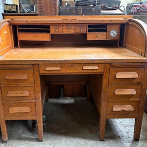 May include: A medium-brown oak roll-top desk featuring a tambour sliding cover, a central drawer, and two pedestals with three drawers each. The interior includes multiple small storage compartments, drawers, and a circular sticker with text.