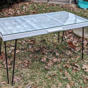 May include: White table with a glass top and a grid of compartments. The table has four black hairpin legs.