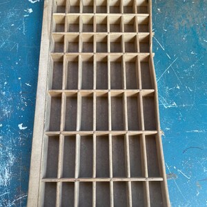 May include: A rectangular wooden printer's drawer featuring a grid of numerous small, open compartments. The light-colored wood frame and dividers create a structured storage layout with a flat, dark-toned base visible within each individual section.