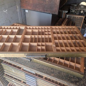 May include: A vintage wooden letterpress type drawer featuring a grid of numerous small rectangular compartments for organizing metal type. The drawer has a weathered olive green metal front edge with a dark handle and a light wood interior.
