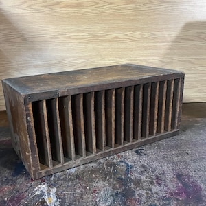 May include: A rectangular wooden storage organizer featuring a series of vertical dividers creating multiple narrow slots. The wood has a dark, weathered brown finish with visible grain and signs of wear across the top and side surfaces.