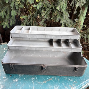 May include: A dark grey metal cantilever tool box featuring two tiered, light grey interior trays with multiple compartments for organisation. The exterior base is a solid dark grey with three metal latches along the front edge for secure closure.