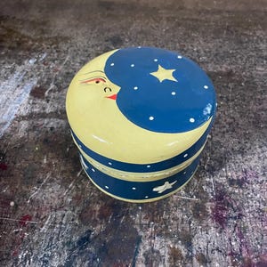 May include: A round wooden trinket box featuring a painted yellow crescent moon face on a dark blue background with white polka dots and a yellow star on the lid. The sides of the box are dark blue with white dots and a small white star accent.