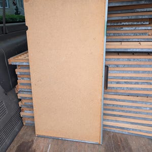May include: A large, rectangular, light brown wooden panel with a metal frame, leaning against a stack of similar panels. The panels appear to be part of a larger structure or furniture piece, possibly for storage or display.