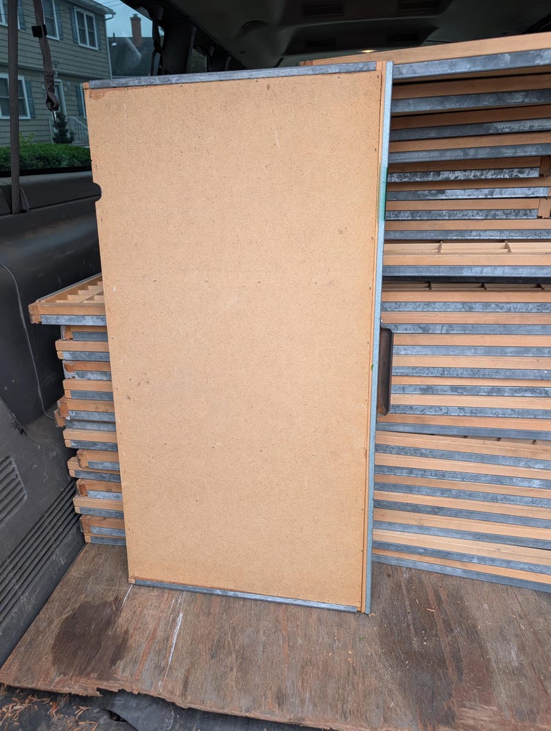 May include: A large, rectangular, light brown wooden panel with a metal frame, leaning against a stack of similar panels. The panels appear to be part of a larger structure or furniture piece, possibly for storage or display.