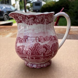 May include: A ceramic pitcher featuring a red and white transferware pattern. The design depicts a landscape with a large manor house, trees, and a body of water. The rim and base have a scalloped edge, and the upper border displays floral motifs.