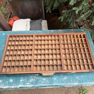 May include: A rectangular wooden printer's drawer featuring a grid of numerous small, open compartments arranged in three distinct sections. The tray is constructed from brown wood and includes a small metal handle centered on the front edge.