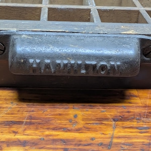 May include: Close-up of a vintage wooden printer's tray with a dark metal handle. The handle is inscribed with the word "HAMILTON" and is attached with screws. The tray has multiple small compartments.