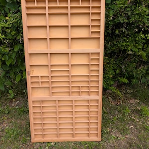 May include: A rectangular light-colored wooden display case featuring numerous small, varied compartments arranged in a grid pattern. The structure includes vertical and horizontal dividers creating shelves of different sizes for organizing small items.