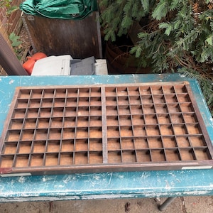May include: A rectangular wooden printer's tray featuring a grid of numerous small, open compartments. The tray is constructed from dark brown wood and is divided into two main sections by a central vertical divider, creating a uniform storage layout.