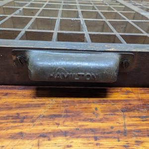 May include: A vintage wooden printer's tray with numerous small compartments. The tray has a dark finish and a metal handle with the name "HAMILTON" embossed on it. The tray is set on a wooden surface.