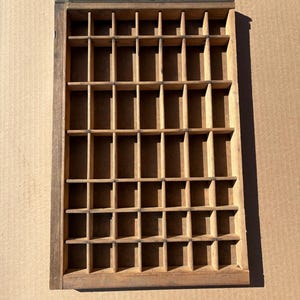 May include: A rectangular wooden printer's tray featuring a grid of numerous small, open compartments. The tray is constructed from light brown wood with visible grain and has a dark metal handle attached to the top edge of the frame.