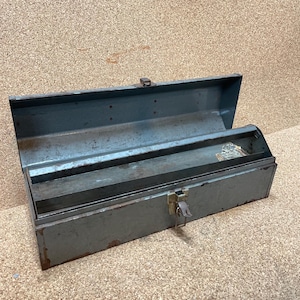 May include: A rectangular gray metal tool box with an open hinged lid and a central metal latch. The box features a weathered finish with visible signs of rust along the bottom edges and interior surfaces, including a long internal tray.