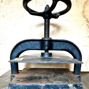 May include: A black metal book press with a large hand crank. The press has two flat plates and a screw mechanism. The press is on a wooden surface.