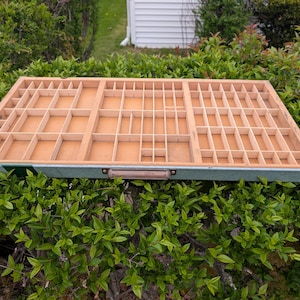 May include: A rectangular wooden printer's tray with numerous small compartments. The tray has a light brown interior and a green and brown border. A small handle is attached to the front. The tray is set against a backdrop of green foliage.