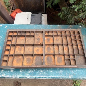 May include: A rectangular wooden printer's type case featuring a grid of numerous small, open compartments of varying sizes. The wood has a dark brown finish with visible signs of wear, including patches of lighter, worn-out areas on the base.