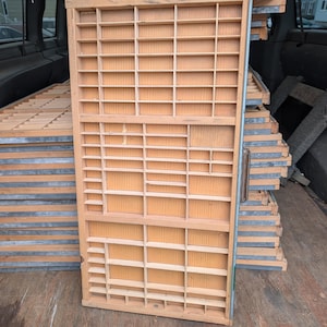 May include: A stack of vintage wooden printer's trays with numerous small compartments. The trays are made of light-colored wood and have metal edging. Several trays are stacked together, suggesting a collection or storage of items.