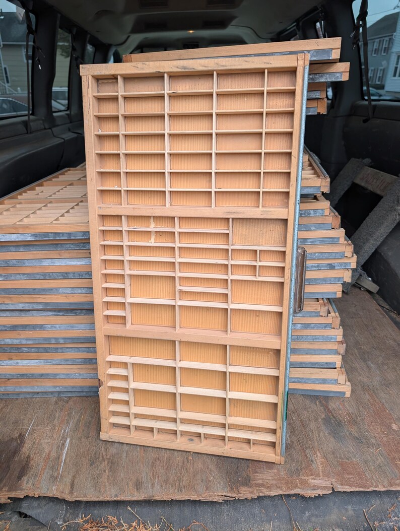 May include: A stack of vintage wooden printer's trays with numerous small compartments. The trays are made of light-colored wood and have metal edging. Several trays are stacked together, suggesting a collection or storage of items.