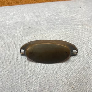 May include: A semi-oval, cup-style drawer pull made of dark, aged brass with a smooth, rounded surface. The hardware features two small, circular mounting holes on either side for attachment, showcasing a weathered, metallic patina finish.