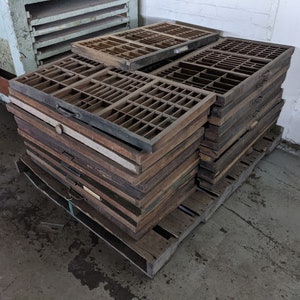May include: A stack of vintage wooden printer's trays with multiple small compartments. The trays are various shades of brown and are stacked on a wooden pallet. Some trays have handles.