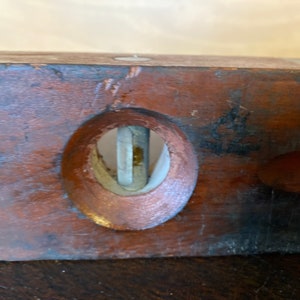 May include: Close-up of a wooden level with a metal bubble vial. The level is brown and has a worn finish. The bubble vial is clear and has a metal frame.