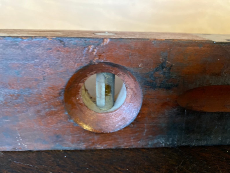 May include: Close-up of a wooden level with a metal bubble vial. The level is brown and has a worn finish. The bubble vial is clear and has a metal frame.