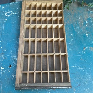 May include: A rectangular wooden printer's tray featuring a grid of numerous small, open compartments. The light-colored wood frame surrounds a series of vertical and horizontal dividers that create a structured storage layout for small items.