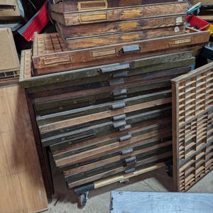 May include: A stack of vintage wooden printer's type drawers with metal handles and label holders. The drawers are various shades of brown and green, some with visible numbers. The drawers are on wheels.