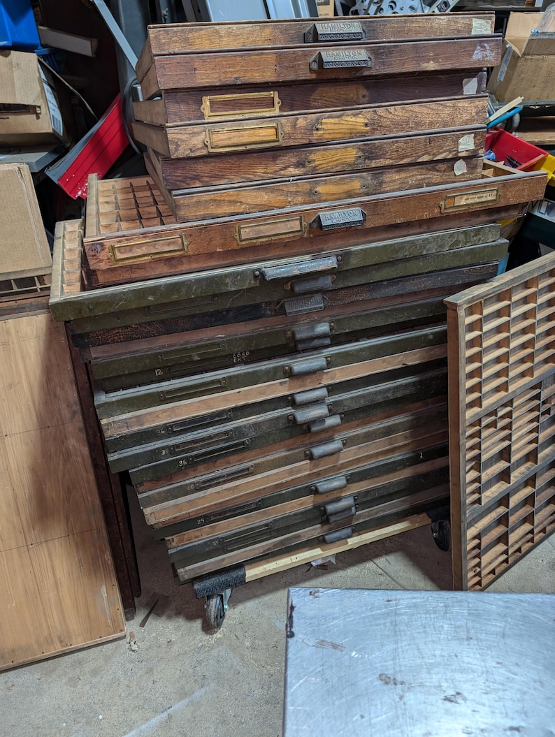 May include: A stack of vintage wooden printer's type drawers with metal handles and label holders. The drawers are various shades of brown and green, some with visible numbers. The drawers are on wheels.