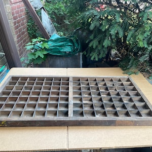 May include: A rectangular wooden printer's tray featuring numerous small, square compartments arranged in a grid pattern. The dark brown wood shows signs of age and wear, and the number 25 is written in yellow ink on the front edge of the frame.