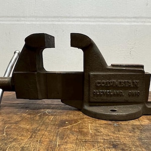 Puede incluir: Un tornillo de banco de hierro fundido vintage con acabado negro. El tornillo de banco tiene una sola manija y una mordaza fija. El tornillo de banco está etiquetado como "Columbian Cleveland, Ohio".
