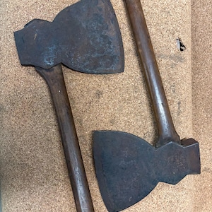 Può includere: Due accette vintage con manici in legno marrone scuro e teste in metallo ossidato e usurato. L'accetta a sinistra presenta un piccolo adesivo rettangolare bianco con il testo 627901 1 vicino alla base del manico. Entrambi mostrano segni d'uso.