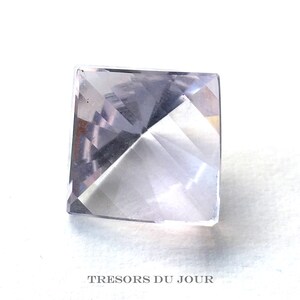 May include: A clear, faceted, square-shaped gemstone with a pale lavender hue. The gemstone is cut into a pyramid shape with a pointed top.