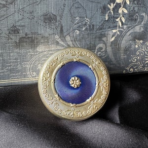 May include: A round, gold-coloured compact with a blue enamel centre. The compact is decorated with a floral design around the edge.