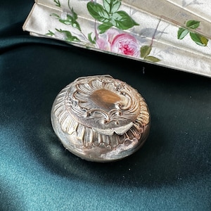 May include: A small, round, silver-coloured compact with ornate detailing on the lid. The compact sits on a dark green surface, with a partially visible, folded fan in the background, decorated with pink roses and green leaves.