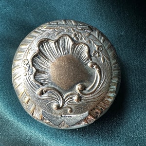May include: A circular, antique silver compact with an elaborate, embossed design. The central feature is a stylized shell shape, encircled by floral and scrollwork details. The compact has a tarnished, aged appearance.