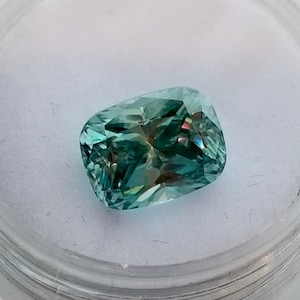 May include: A teal-coloured cushion-cut gemstone with iridescent flashes, set in a clear plastic container with a white interior. The gem's facets reflect light, creating a sparkling effect.